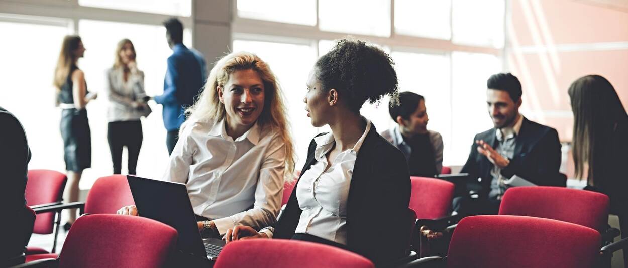 Foto van een conferentiezaal: midden in beeld zitten twee vrouwen op stoelen te praten. Achter hen zit nog een groepje praters en helemaal achter in beeld staat een groepje aan de zijkant van de zaal.