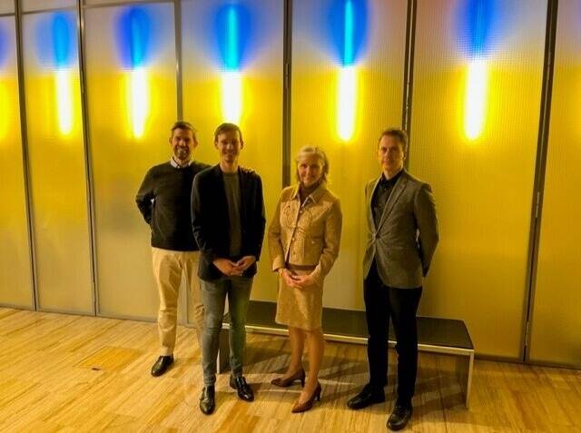 Groepsfoto van drie collega's van de academie en de operationeel manager van de Nederlandse ambassade in Berlijn. Ze staan en kijken recht in de camera met blauwgeel neonlicht op de achtergrond.