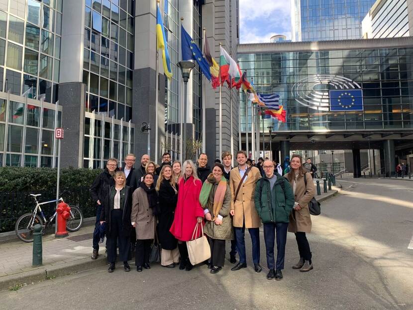 Groepsfoto deelnemers training EU Historisch Perspectief (DGES Masterclass). Ze staan voor het gebouw van de Europese Unie in Brussel en op de achtergrond zie je vlaggen wapperen.
