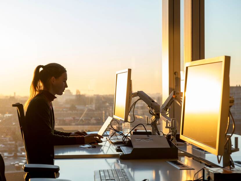 Woman working behind a desk with two screens. She's sitting before a big window and the sun is setting.